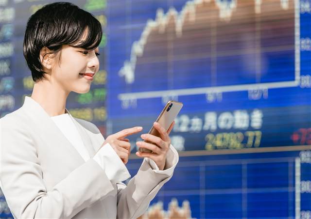 株価ボードの前でスマホを見る読者のイメージ